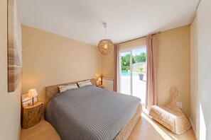 Un dormitorio con una cama grande, una mesita de noche con lámpara, una silla de mimbre y vistas al exterior a través de una puerta corredera.