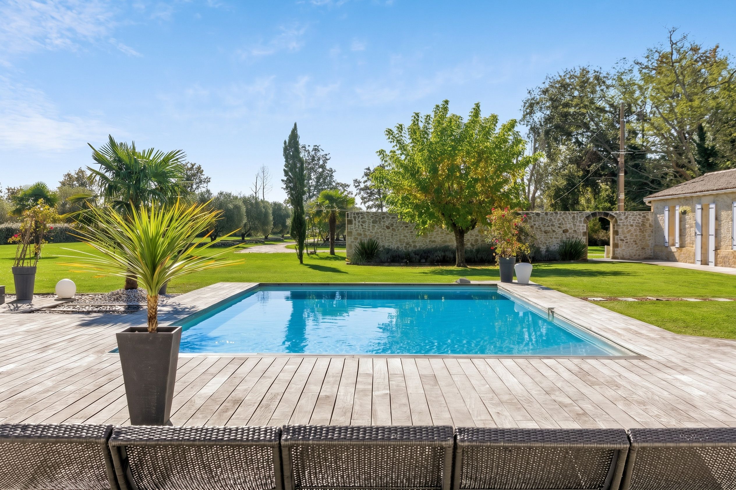 A rectangular swimming pool with a wooden deck surrounding it, a stone wall, and a building with a tiled roof in the background.