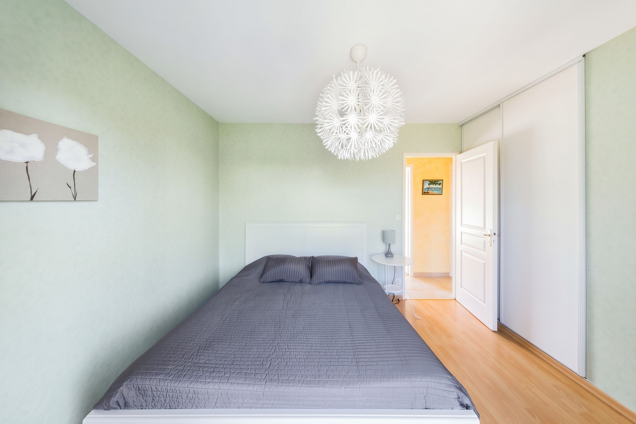 A bedroom with a bed, a white headboard, a wall-mounted lamp, and a door leading to another room.