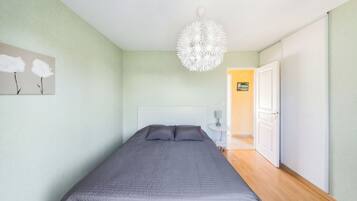 A bedroom with a bed, a white headboard, a wall-mounted lamp, and a door leading to another room.