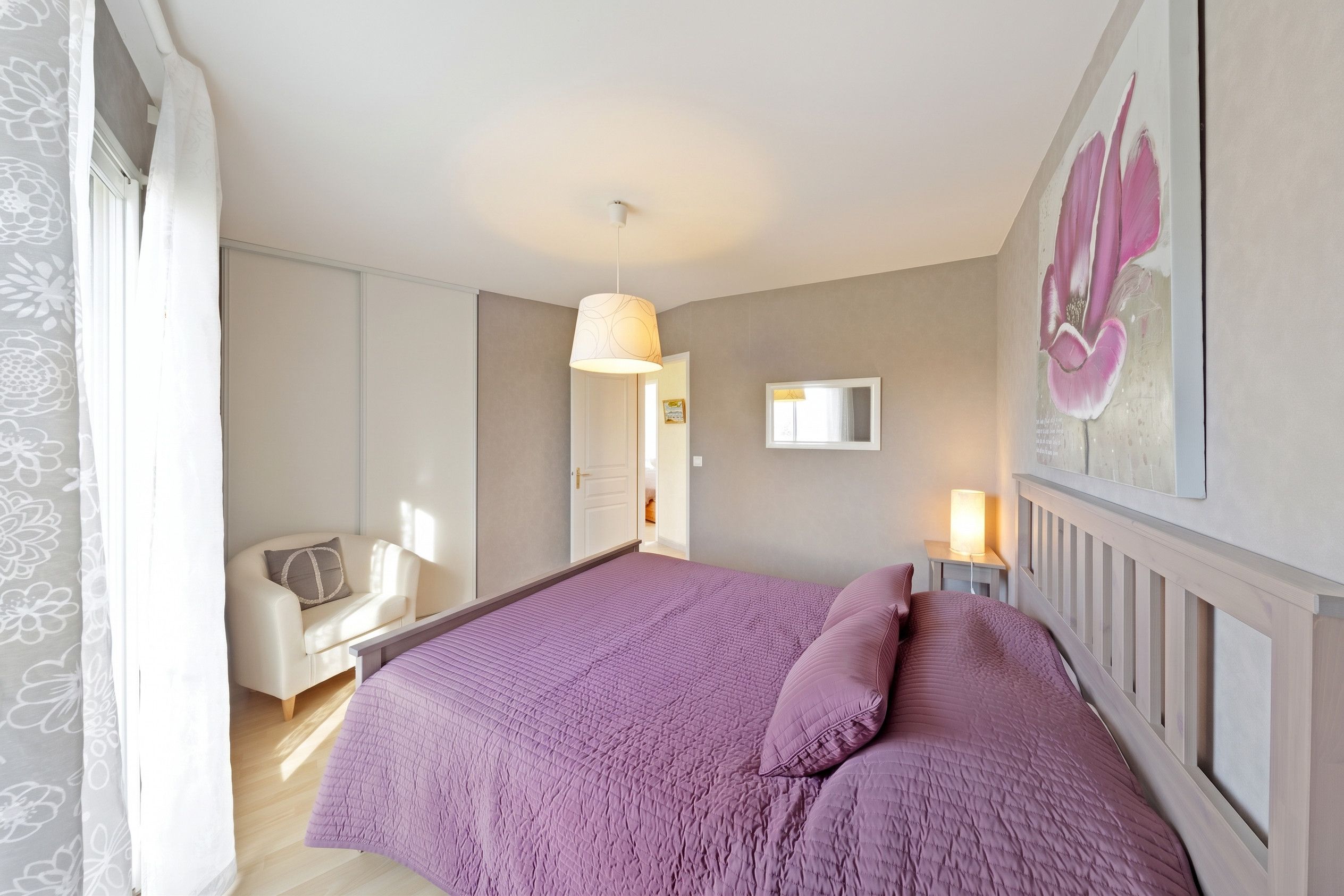 Une chambre à coucher comprenant un lit, une chaise, une table de chevet avec une lampe, un tableau au mur et une porte donnant sur une autre pièce.