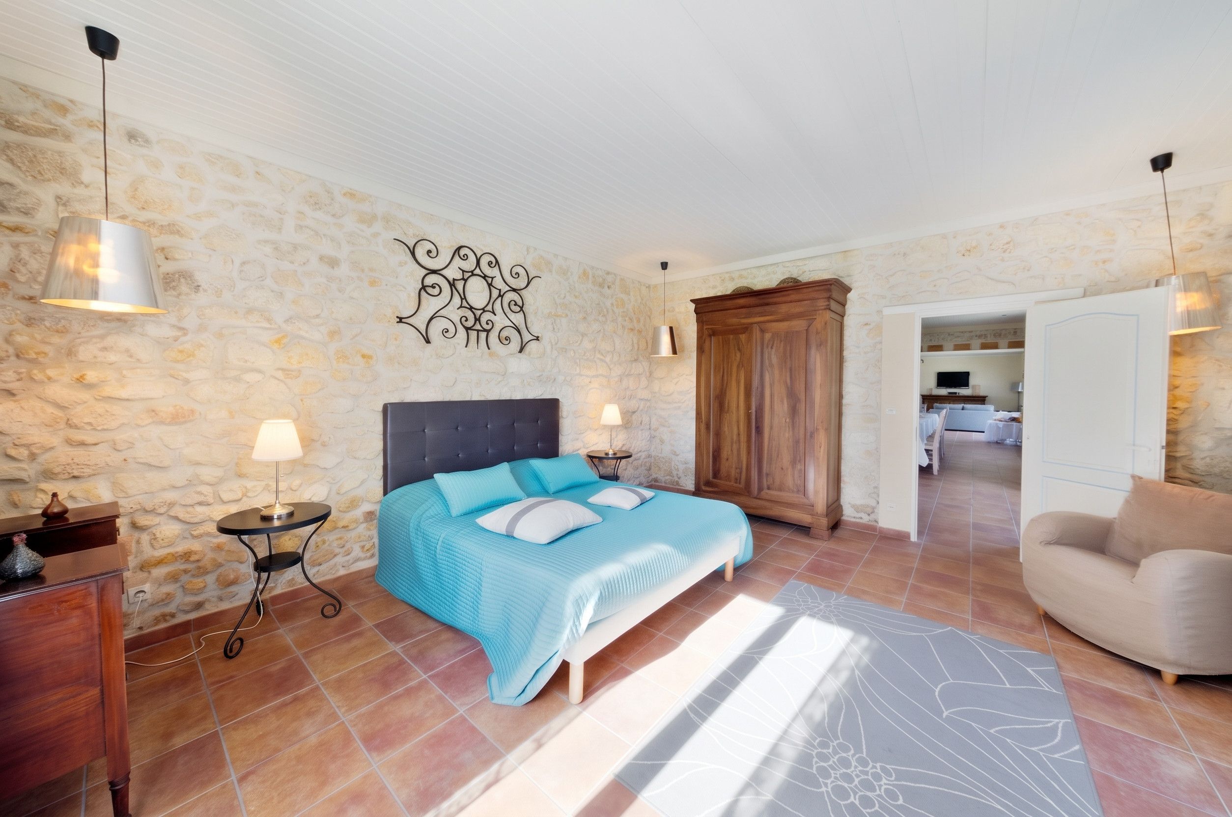 Une chambre avec un mur en pierre, un lit avec une literie bleue, deux lampes de chevet et une armoire en bois.