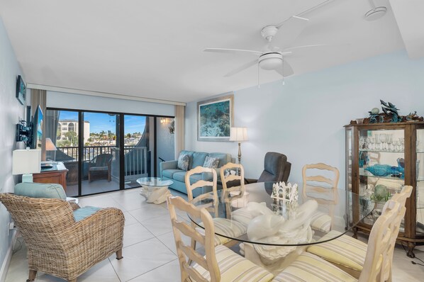 A spacious living room with a balcony, a ceiling fan, a dining table with chairs, and a glass cabinet with decorative items.