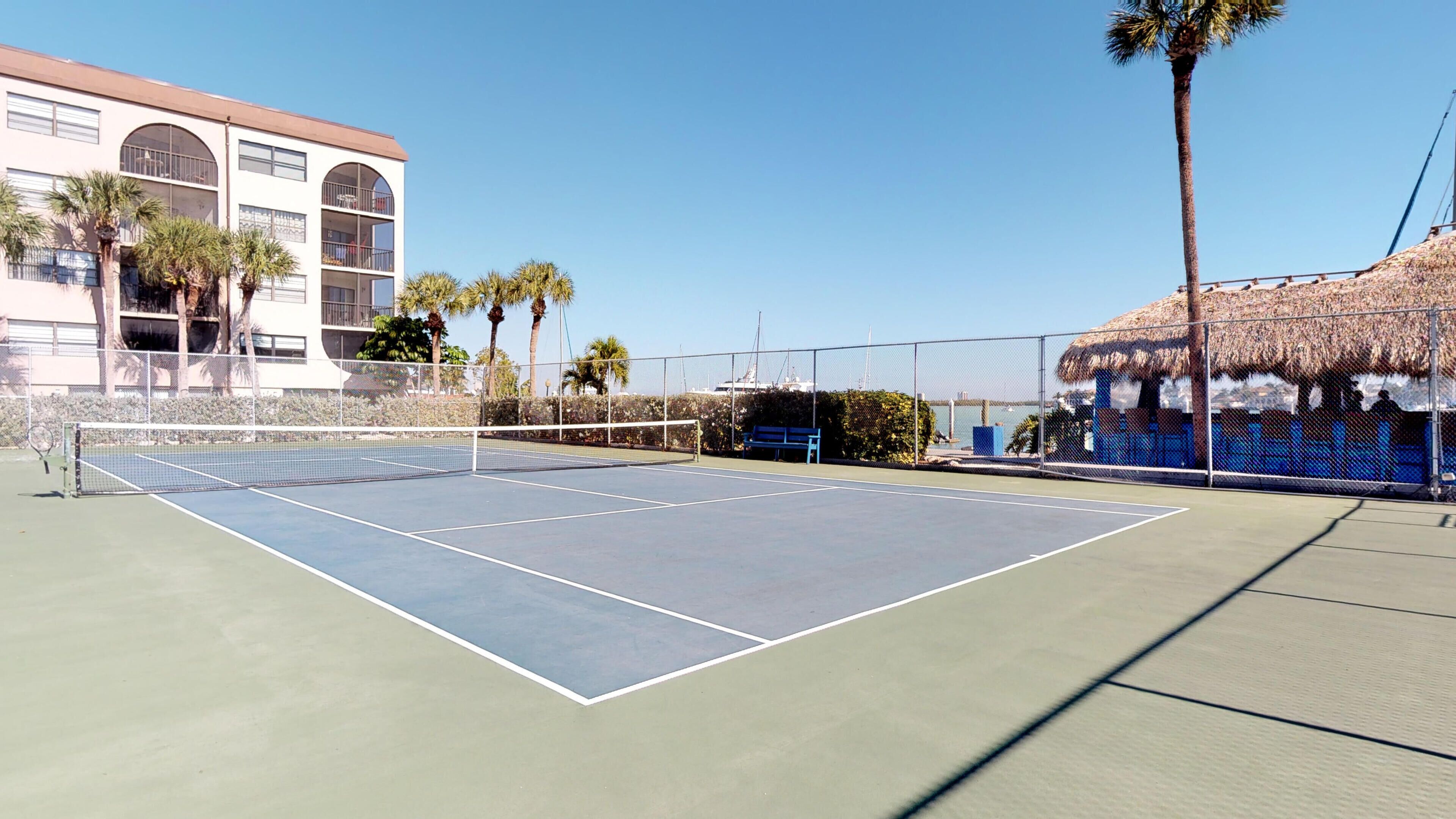 An outdoor tennis court with a net, surrounded by a fence, palm trees, and a building with arched windows.