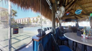 En bar vid stranden med ett halmtak, utomhusservering och utsikt över en tennisbana.