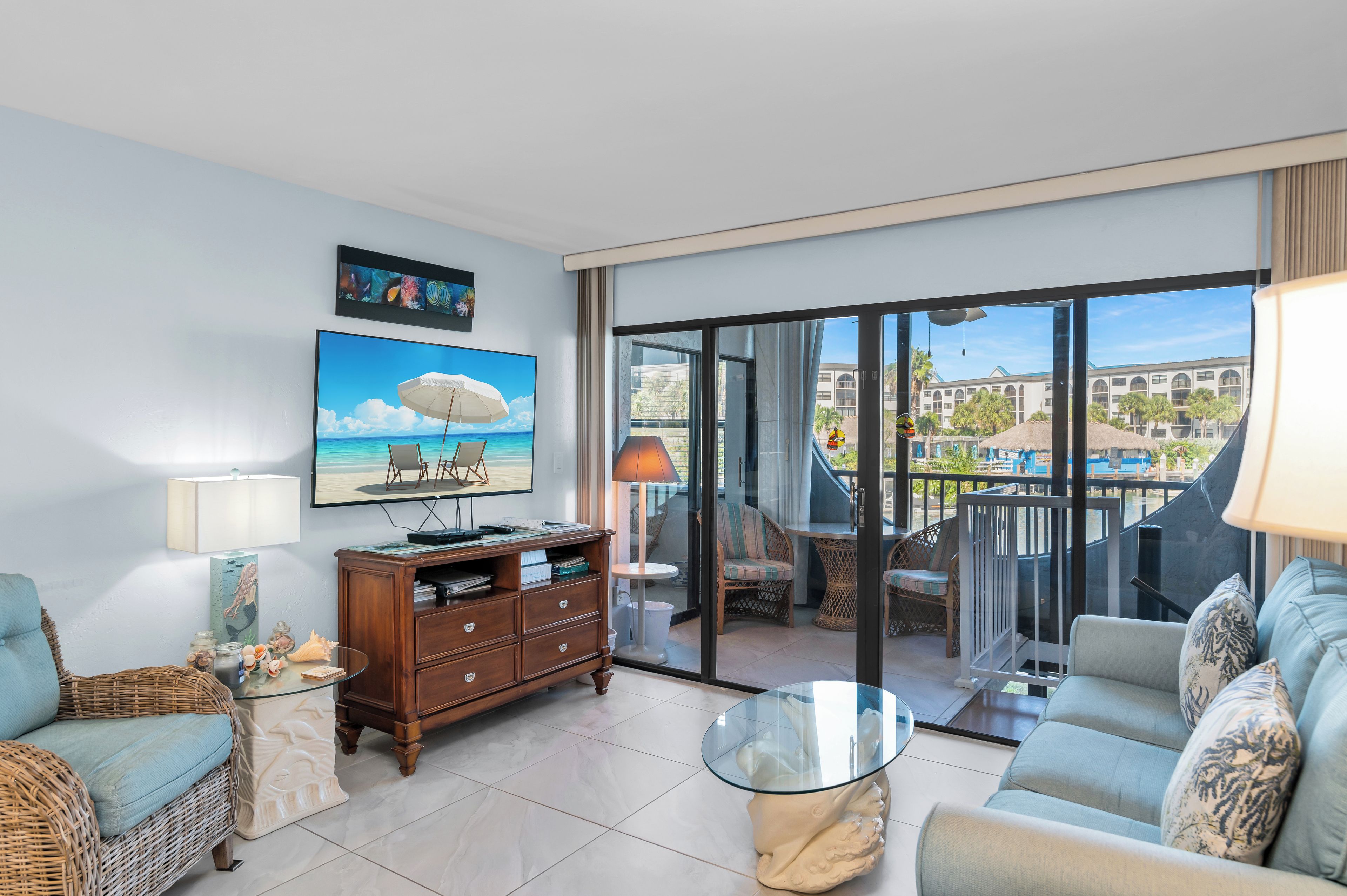 A spacious living room with a large window, a flat-screen TV, a comfortable sofa, and a dining area with wicker furniture.