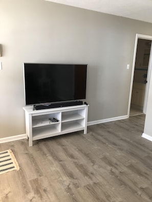 A room with a large flat-screen TV on a white stand, wooden flooring, and a door leading to a kitchen. - Spacious & Close to Beach- Condo E (Ocean City)