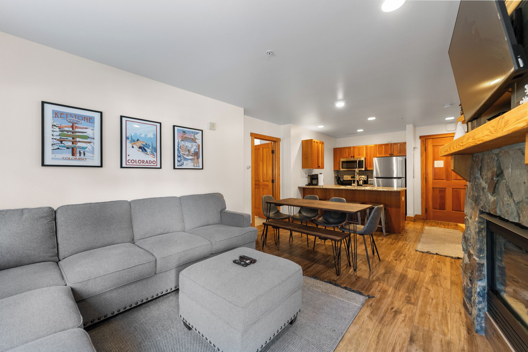 A modern living room with a gray sectional sofa, a small ottoman, and a dining area with a wooden table and chairs. There are framed pictures on the wall, a kitchen with wooden cabinets, and a stone fireplace.