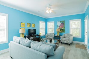 Una sala de estar con una pared azul, un ventilador de techo, un sofá, dos sillones, una mesa de centro, un televisor y cuadros enmarcados.