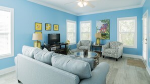 Un salon avec un mur bleu, un ventilateur de plafond, un canapé, deux fauteuils, une table basse, un téléviseur et des tableaux encadrés.