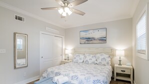 Une chambre à coucher comprenant un lit, un ventilateur de plafond, une table de chevet, une commode, un miroir et un tableau accroché au mur.