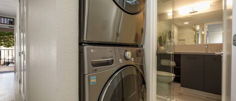 A modern laundry room with stacked washers and dryers, a glass shower enclosure, and a contemporary kitchen with dark cabinetry.