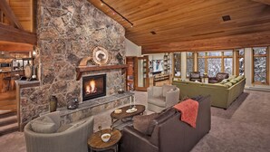 Una sala de estar acogedora con chimenea de piedra, pared de piedra y un ventanal con vistas nevadas.