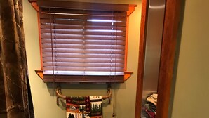 Un pasillo estrecho con una puerta de madera, una ventana con persianas, un estante pequeño con varios objetos y un suelo de baldosas.