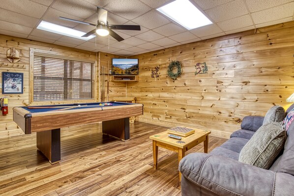 A pool table, a sofa, and a television in a room with wooden walls and ceiling fan.
