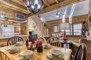 A cozy cabin interior with a wooden interior, a dining area with a table set for four, a staircase, and a Christmas tree.