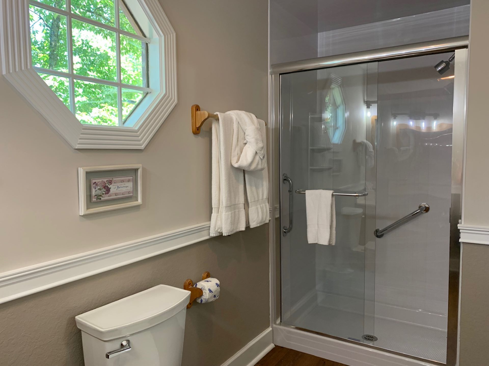 A bathroom with a toilet, a framed picture, a towel rack with towels, and a glass-enclosed shower.