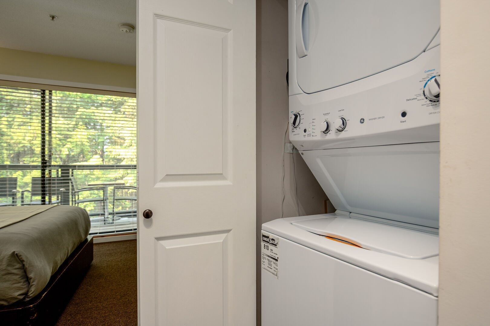 A laundry room with a front-loading washer and dryer, a bed with a beige cover, and a window with blinds overlooking a balcony.