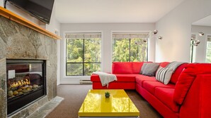 A modern living room with a stone fireplace, a red sectional sofa, and a yellow coffee table. - 2BR | Powderhorn | Near Blackcomb Ski Area | Double Sided Fireplace | Rooftop Hot Tub | Parking (Whistler)