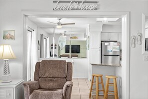 Uma sala moderna com uma poltrona marrom, um espaço de refeições com banquetas, uma cozinha com armários brancos e um ventilador de teto.