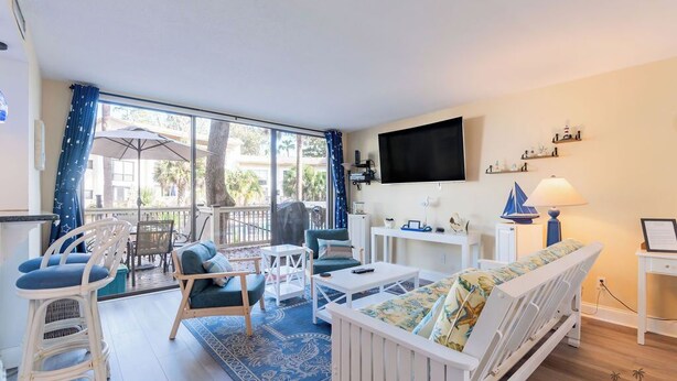 A cozy living room with a sofa, chairs, a coffee table, and a TV. There's a balcony with a view of palm trees and buildings.
