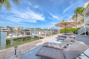 A waterfront patio with lounge chairs, a table with an umbrella, and a dock with boats.