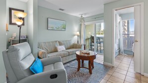 A living room with a sofa, a chair, a table, and a door leading to a balcony. - Easy Beach Days | Studio Near Pool & Shore (Destin)