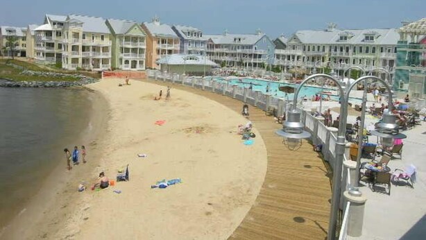 Un hotel frente a la playa con un paseo marítimo de madera, piscinas y gente disfrutando de la playa.
