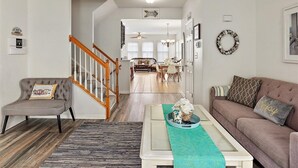 Una sala de estar con un sillón gris, una mesa de centro blanca con un camino de mesa turquesa, un sofá marrón con una almohada que dice 'Enjoy', y una escalera que lleva a otra habitación.
