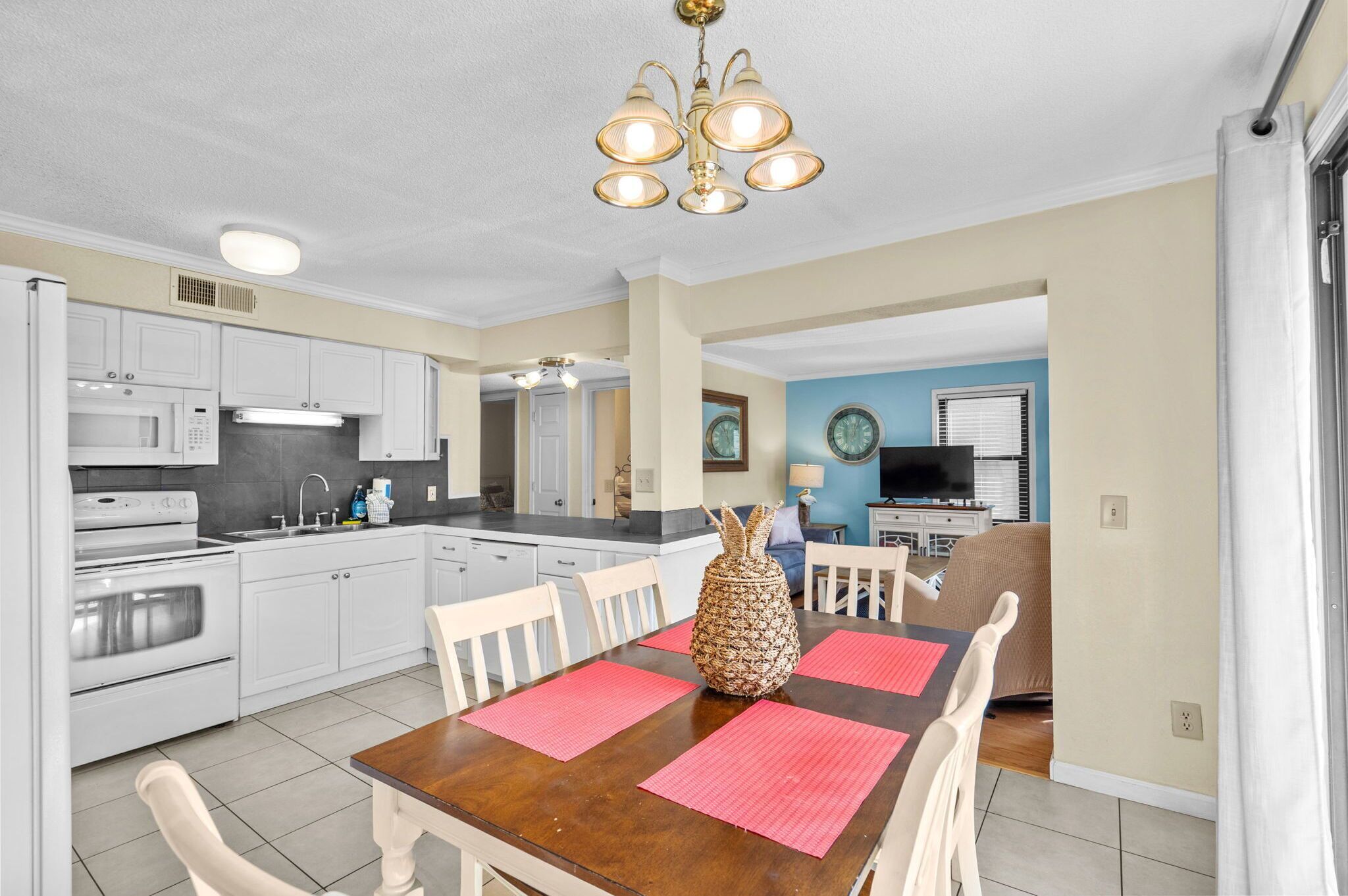 Une cuisine moderne avec des meubles blancs, un four à micro-ondes et une cuisinière. On y trouve une table à manger avec des chaises et un ananas en guise de décoration centrale.