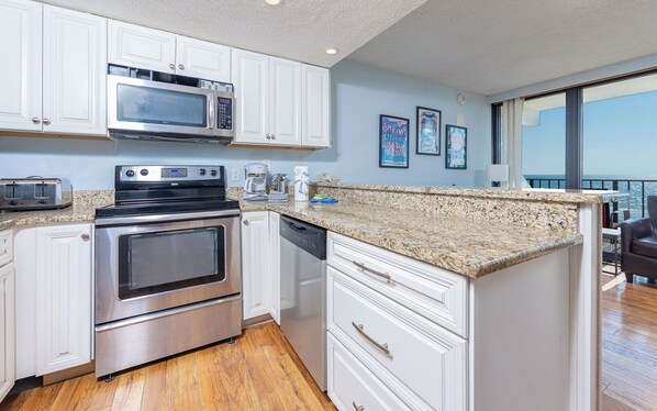 A modern kitchen with white cabinets, stainless steel appliances, a granite countertop, and a view of the ocean through large windows.