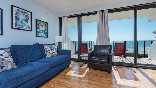 Ein Zimmer mit einem blauen Sofa, einem Ledersessel und einem großen Fenster mit Meerblick.