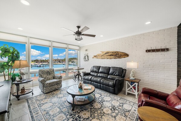 Un salon spacieux doté d’une grande fenêtre, d’un ventilateur de plafond, d’un canapé en cuir, d’un tapis à motifs et d’une vue sur une piscine.
