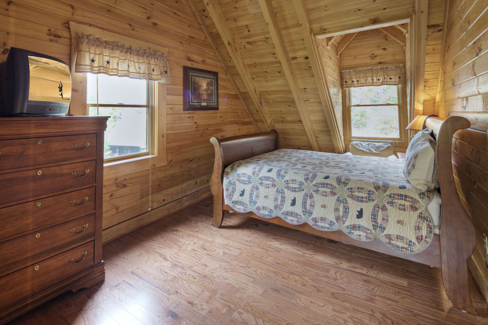 A cozy bedroom with a wooden ceiling, a bed with a patterned quilt, a dresser with drawers, and two windows with curtains.