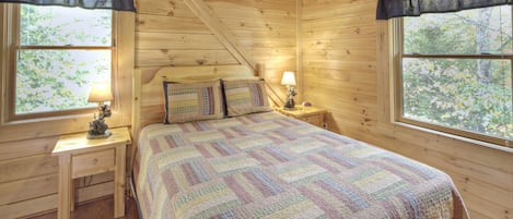 A wooden cabin bedroom with a bed, two bedside tables, and two windows with curtains.