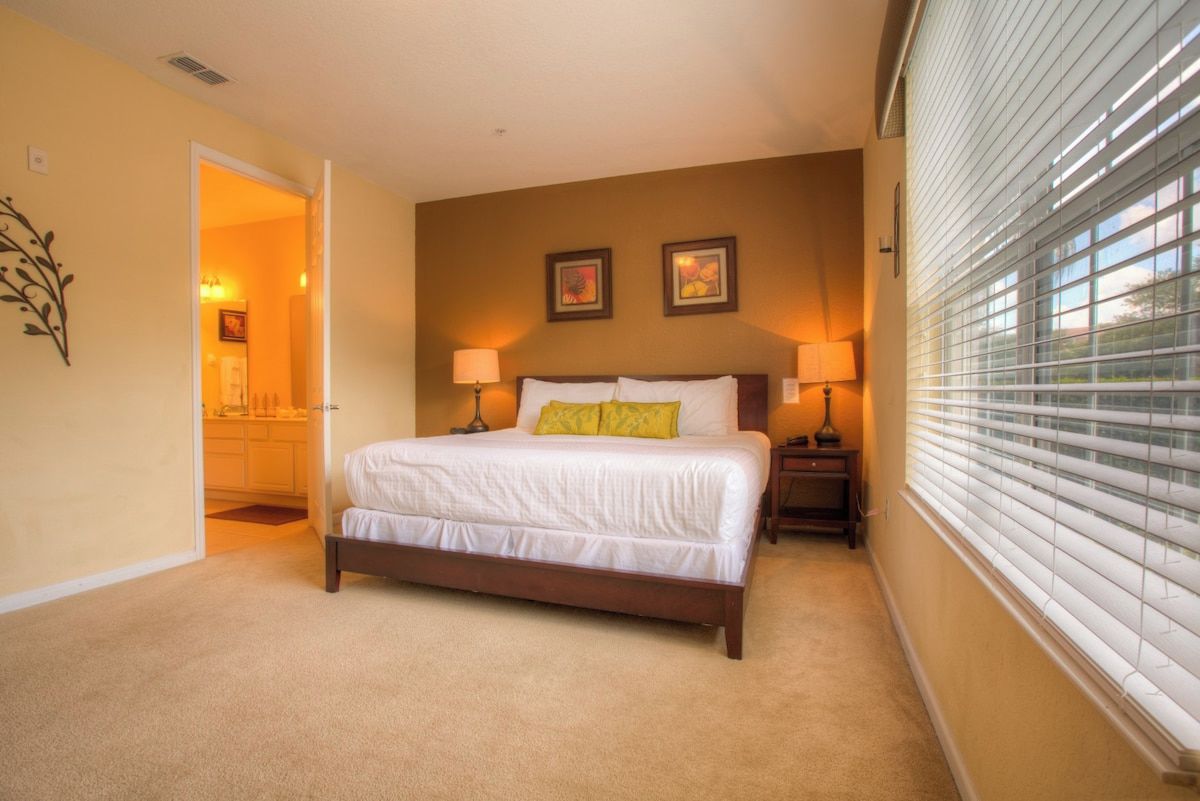 A bedroom with a large bed, two bedside lamps, a window with blinds, and a door leading to a bathroom.