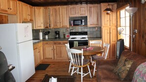 Een kleine keuken met houten kastjes, een witte koelkast, een magnetron en een fornuis. Er staat een eettafel met stoelen en een bruine bank met een decoratief plaid.