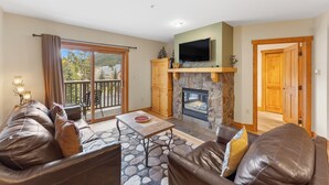 A living room with a stone fireplace, leather sofas, a flat-screen TV, and a balcony with a view of trees and a house.