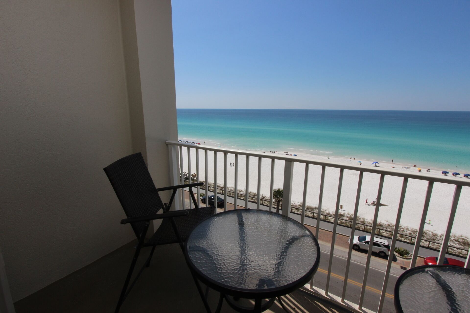A balcony with a glass table and chairs, overlooking a beach with a clear view of the ocean and a road with parked cars.