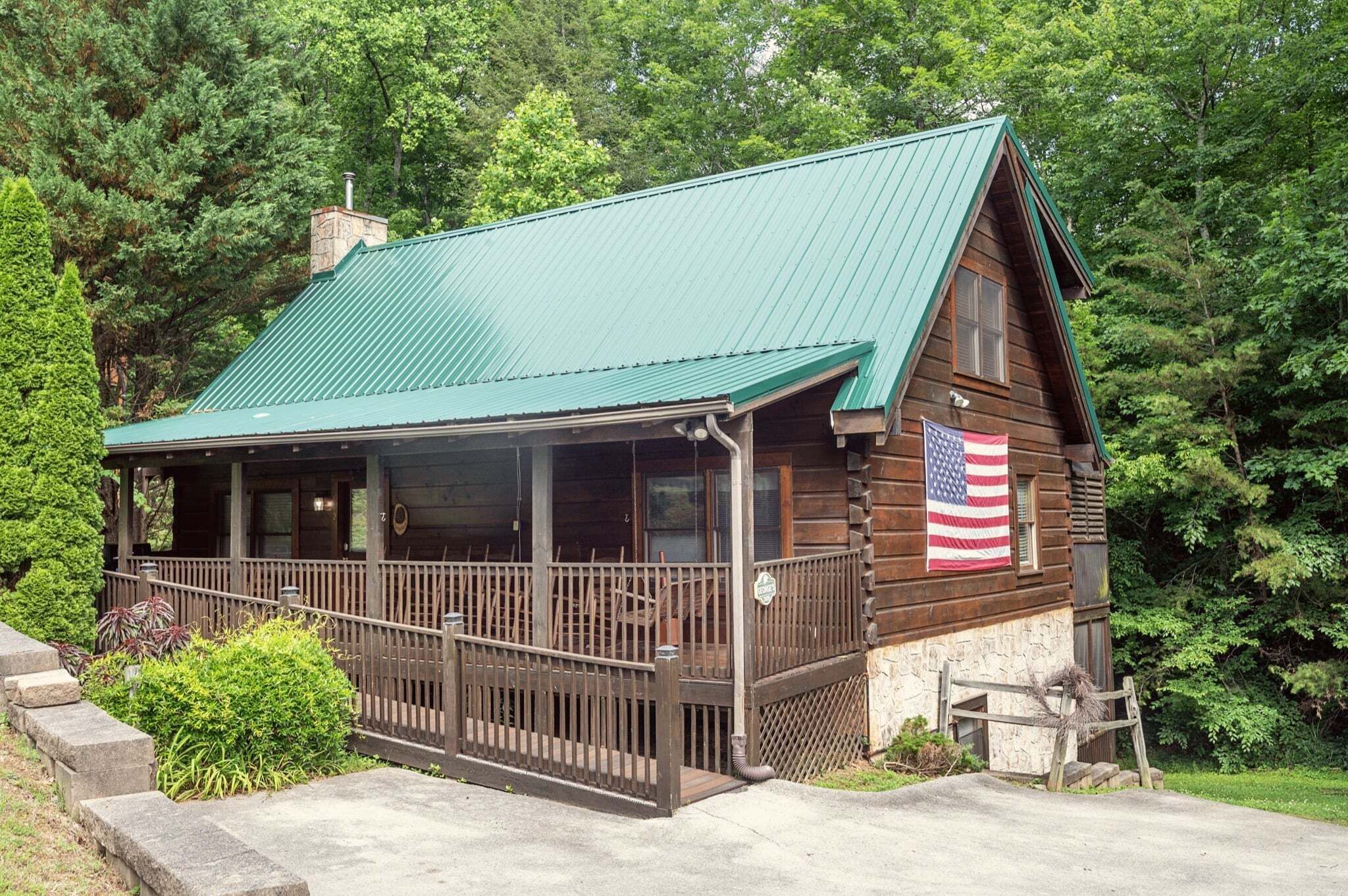 A wooden cabin with a green roof, a porch, and an American flag.