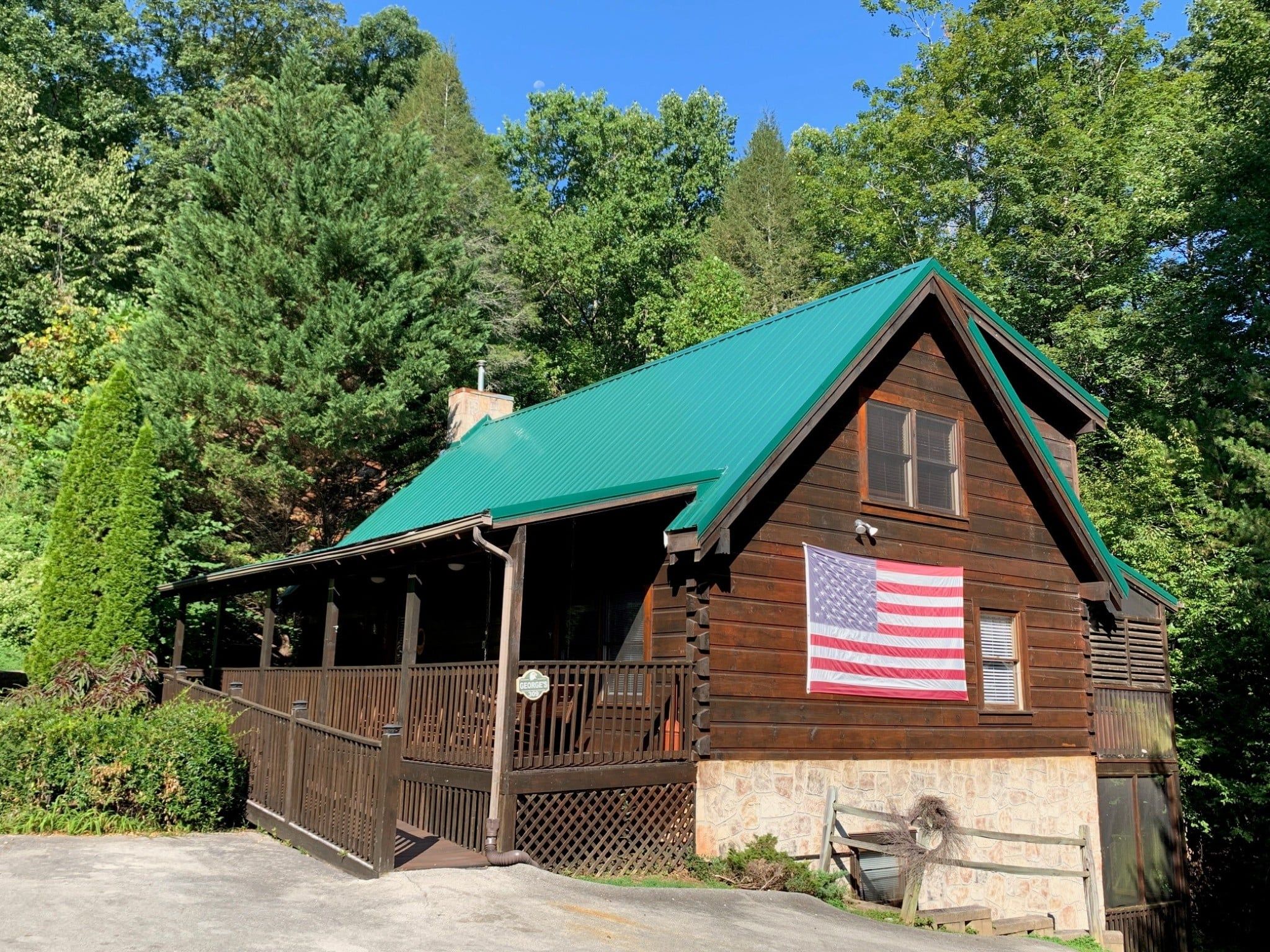 A wooden cabin with a green roof, a porch, and an American flag.