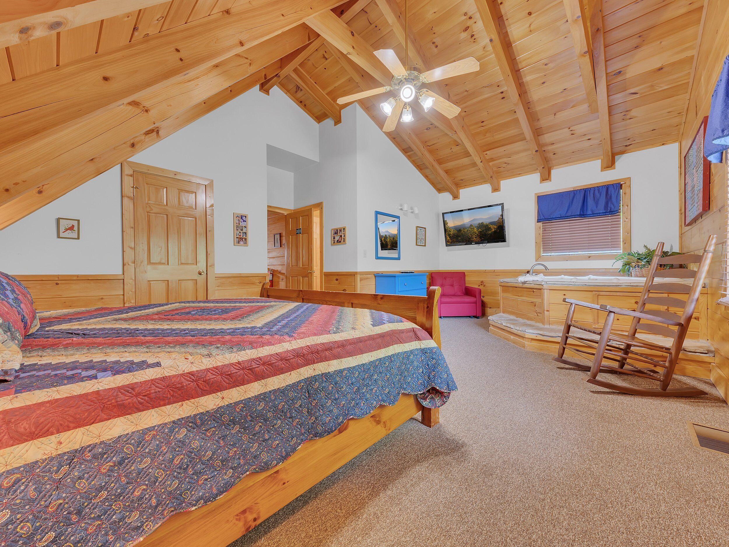 A cozy bedroom with a wooden ceiling, a large bed with a colorful quilt, a pink chair, a wooden rocking chair, and a TV on the wall.