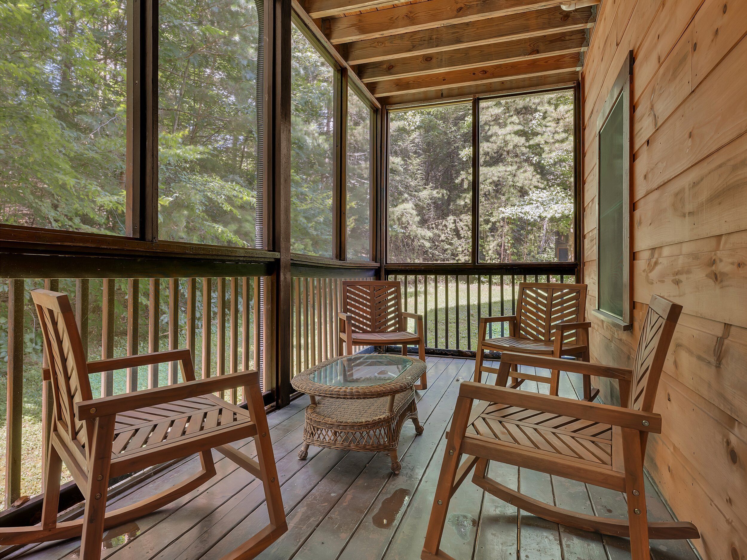 A wooden deck with four wooden chairs and a glass table, surrounded by large windows offering a view of a forest.