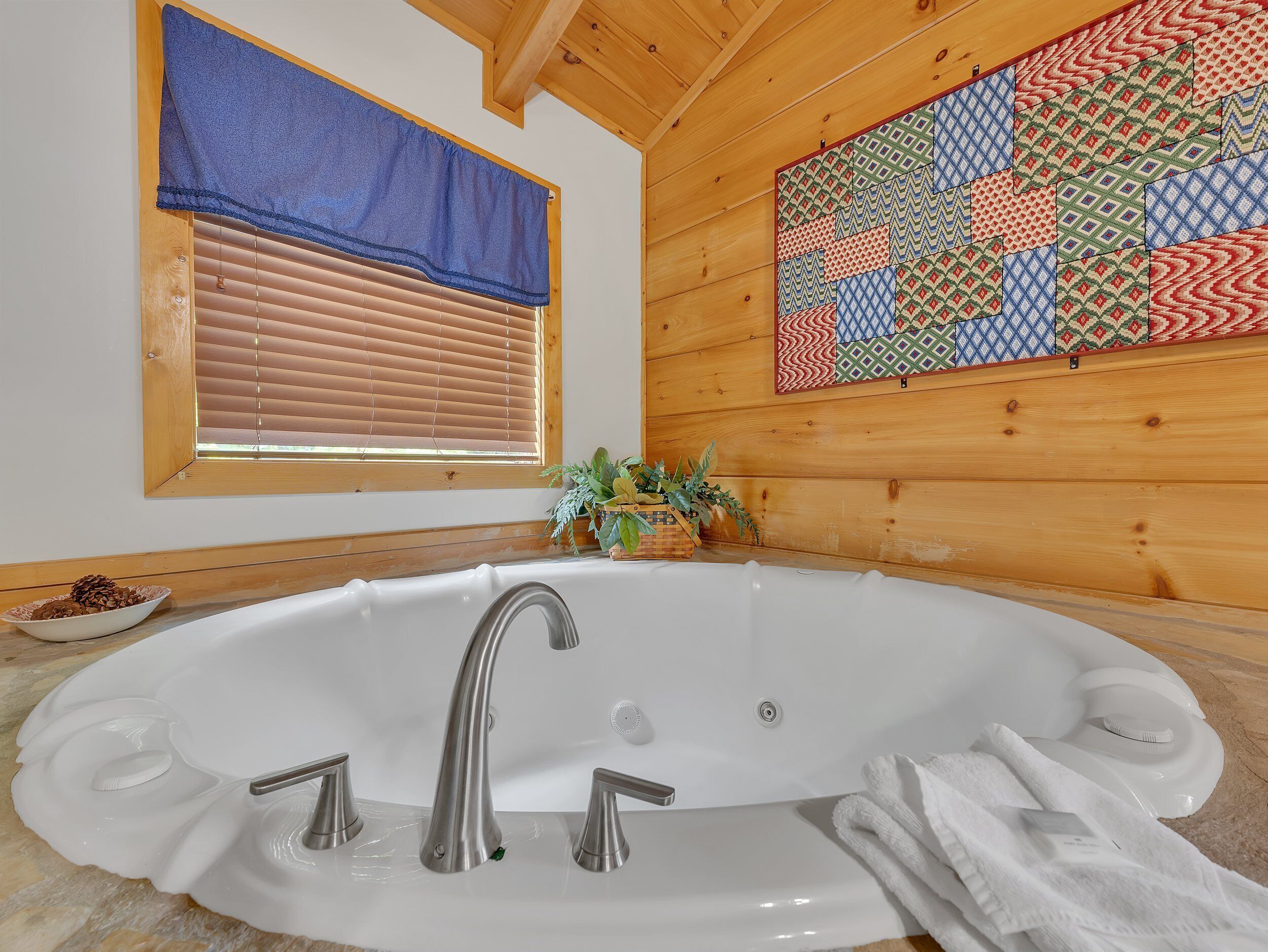Un baño con una bañera independiente, paredes de madera, un ventanal con persianas y un colcha colorida colgada en la pared.
