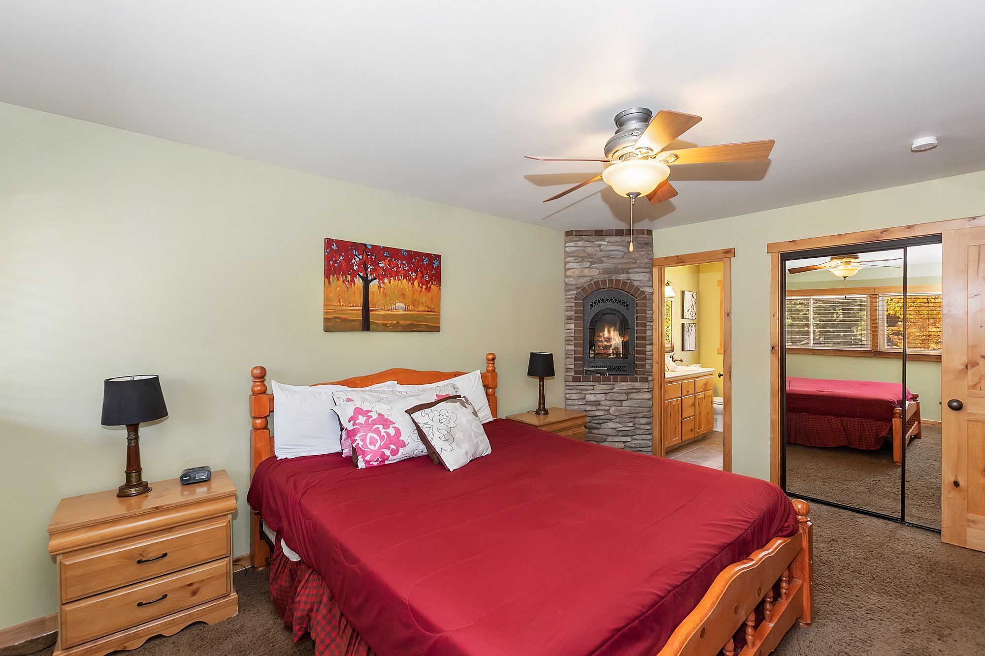 A bedroom with a bed, a nightstand, a lamp, a ceiling fan, a fireplace, and a door leading to another room.