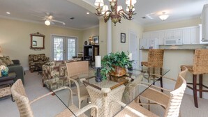 A spacious living room with a glass dining table surrounded by wicker chairs, a chandelier, and a television in the background. - Oceanfront Opulence: Vacation Home with Amazing Pool, Beach Just Moments Away (Pensacola)