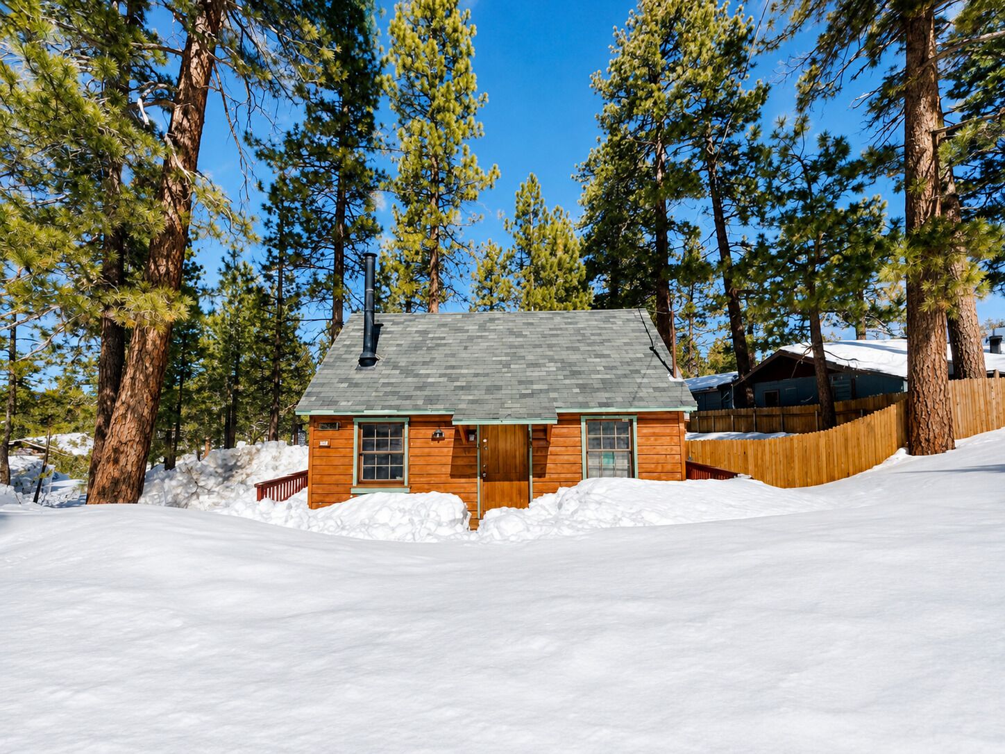 Una cabaña rodeada de altos pinos, con una valla de madera y nieve en el suelo.