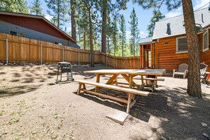 Ein rustikales Blockhaus mit Holzterrasse, Picknicktisch und Sitzbereich im Freien, umgeben von hohen Kiefern.