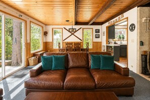 A spacious living room with a brown leather sofa, teal cushions, wooden beams, large windows, and a kitchen visible through an open door.