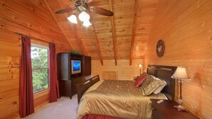 A wooden cabin bedroom with a bed, dresser, TV, and a ceiling fan.